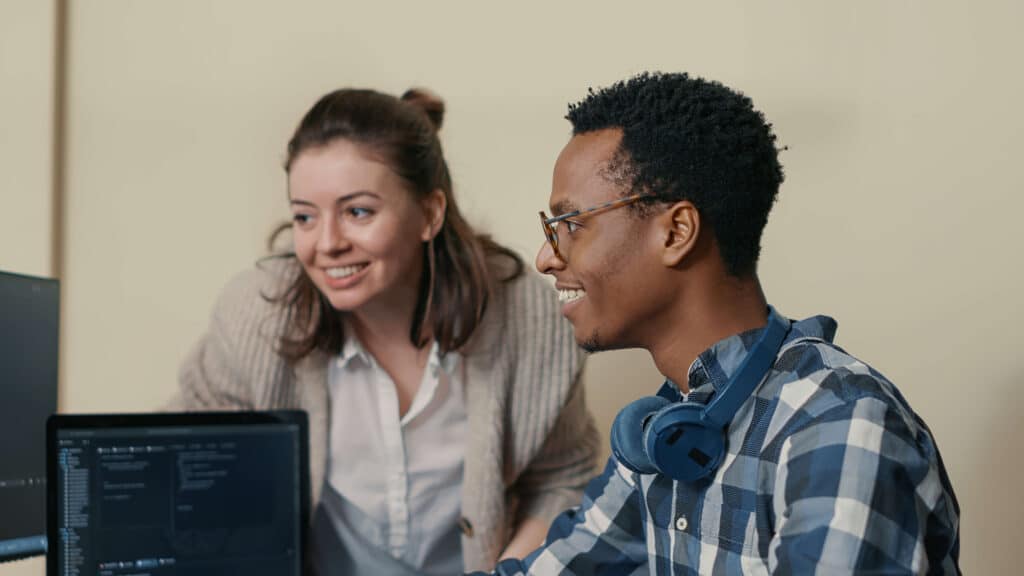 Duas pessoas colaboram em um espaço de escritório. Uma está sentada, vestindo uma camisa xadrez e fones de ouvido no pescoço, trabalhando em um computador para emitir NFe via API. A outra está de pé, vestindo um cardigan leve, sorrindo e olhando para a tela. Ambas parecem engajadas e felizes.