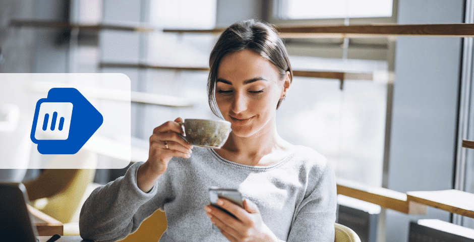 Uma mulher empresária, pele branca, sentada de frente para seu notebook, tomando um café e checando dados em dashboards em seu smartphone