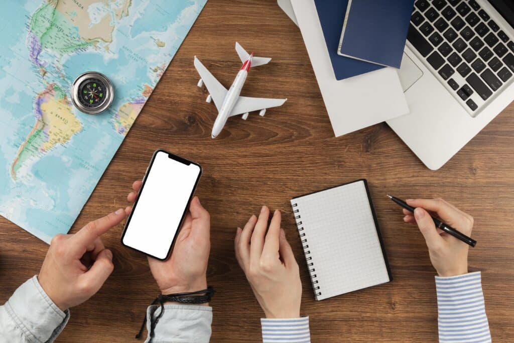 Uma mesa com um mapa, uma bússola, um avião de brinquedo, dois passaportes e um laptop com sistema operacional CTe OS, além de um telefone com tela em branco e um bloco de notas com uma pessoa segurando uma caneta.