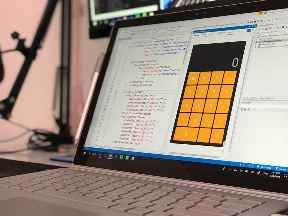 Uma tela de laptop exibe codificação no Visual Studio com uma prévia de um aplicativo de calculadora digital, enquanto ferramentas para Consulta Serasa aparecem no espaço de trabalho ao lado de parte de um microfone.