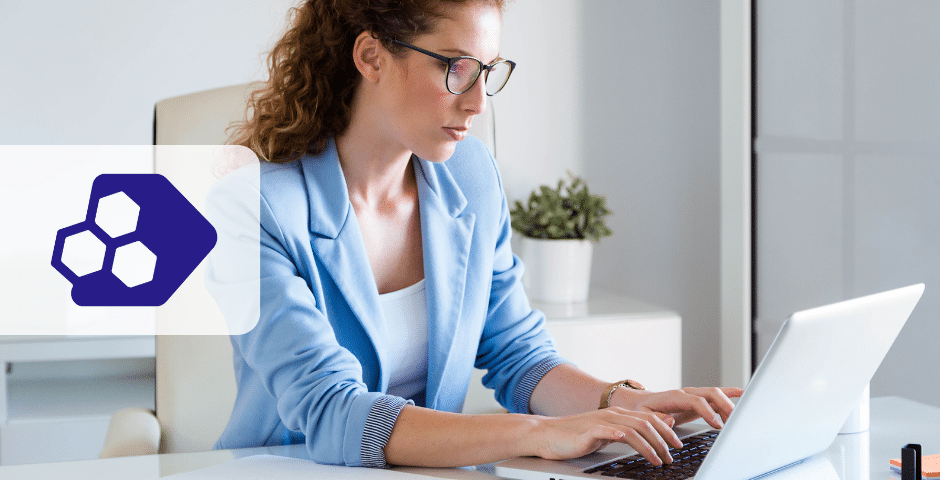 Uma mulher com cabelos cacheados e óculos, vestindo um blazer azul claro, está trabalhando em um laptop em um escritório moderno, sugerindo envolvimento com NFC-e no SPED Fiscal.