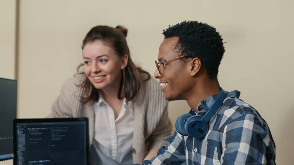 Um homem e uma mulher colaboram em uma mesa com um laptop. O homem, com fones de ouvido e uma camisa xadrez, foca na tela enquanto eles se aprofundam em uma API financeira. Ao lado dele, a mulher de suéter está sorrindo, intrigada com os insights que se desdobram no monitor.
