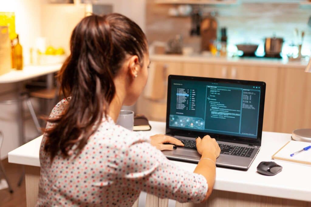 Uma mulher com um rabo de cavalo está sentada em um balcão de cozinha suavemente iluminado, digitando código XML em seu laptop. Vários itens de cozinha criam um cenário convidativo, enquanto um mouse, uma xícara e um bloco de notas para anotar notas de conversão de XML para PDF.