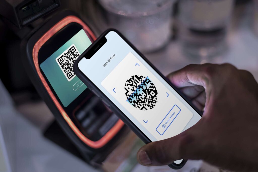 Uma pessoa segura um smartphone com um código QR em frente a um terminal de pagamento, escaneando-o para uma transação digital usando o Pix na Nota Fiscal. Tanto a tela do celular quanto o terminal exibem códigos QR semelhantes.