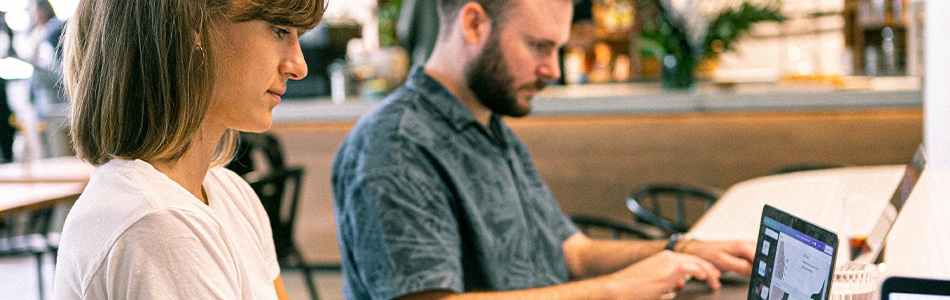 Duas pessoas sentam-se à mesa, trabalhando em laptops em um ambiente claro e casual. A pessoa da esquerda veste uma camisa branca e a da direita ostenta uma camisa estampada. Elas discutem estruturas e como utilizá-los em um cenário de plantas e decoração de interiores desfocadas.