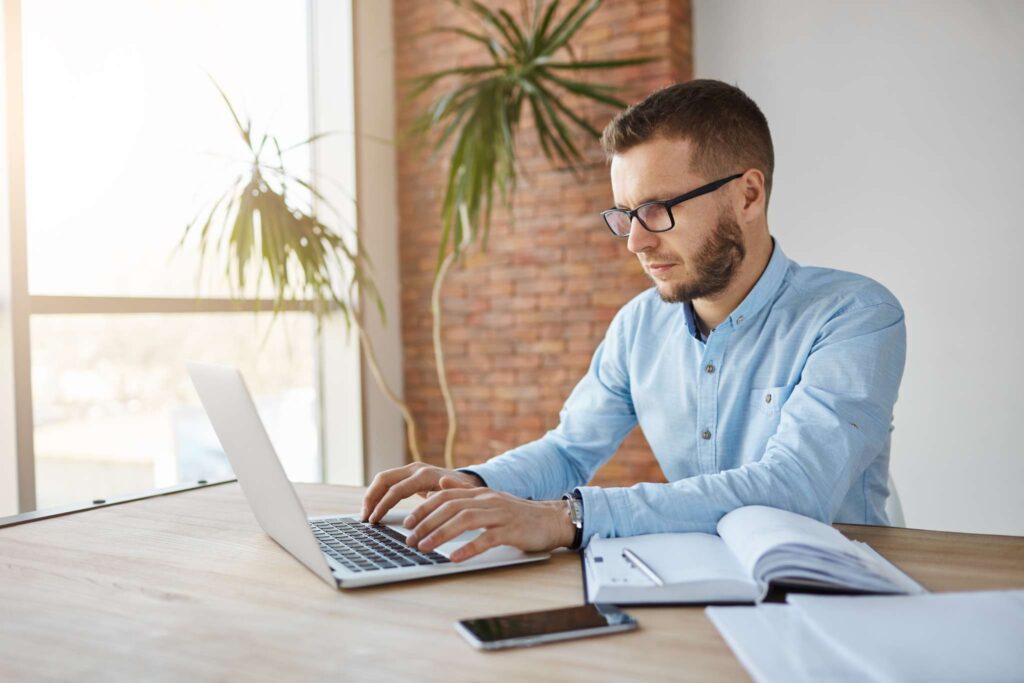 Um homem de óculos e camisa azul-claro trabalha em um laptop em uma mesa com um bloco de notas aberto, caneta, smartphone e ferramentas para armazenamento de documentos fiscais. Uma grande planta e uma parede de tijolos aparecem ao fundo, enquanto a luz do sol entra por uma janela.