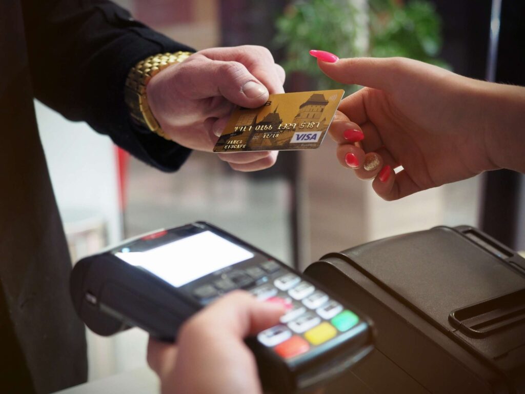 Uma pessoa entrega um cartão de crédito Visa ouro para outra pessoa, que segura uma moeda, perto de um terminal de pagamento com cartão, enquanto uma terceira pessoa, seguindo os padrões de pagamento da FEBRABAN, se prepara para processar a transação.