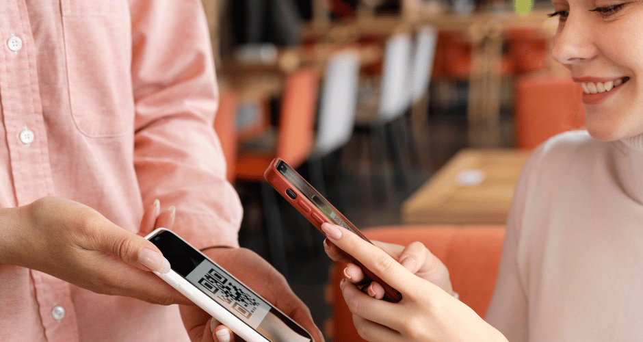 duas mulheres de roupas cor de rosa claro, realizando pagamento por pix, de um celular para o outro, um lendo o qr code pix do outro celular.