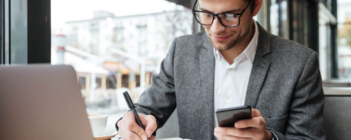 Um homem de óculos e terno senta-se em uma mesa com um laptop, concentrado em Registros SPED Contribuições, segurando um smartphone em uma das mãos e escrevendo em um caderno com a outra, totalmente envolvido em seu trabalho.