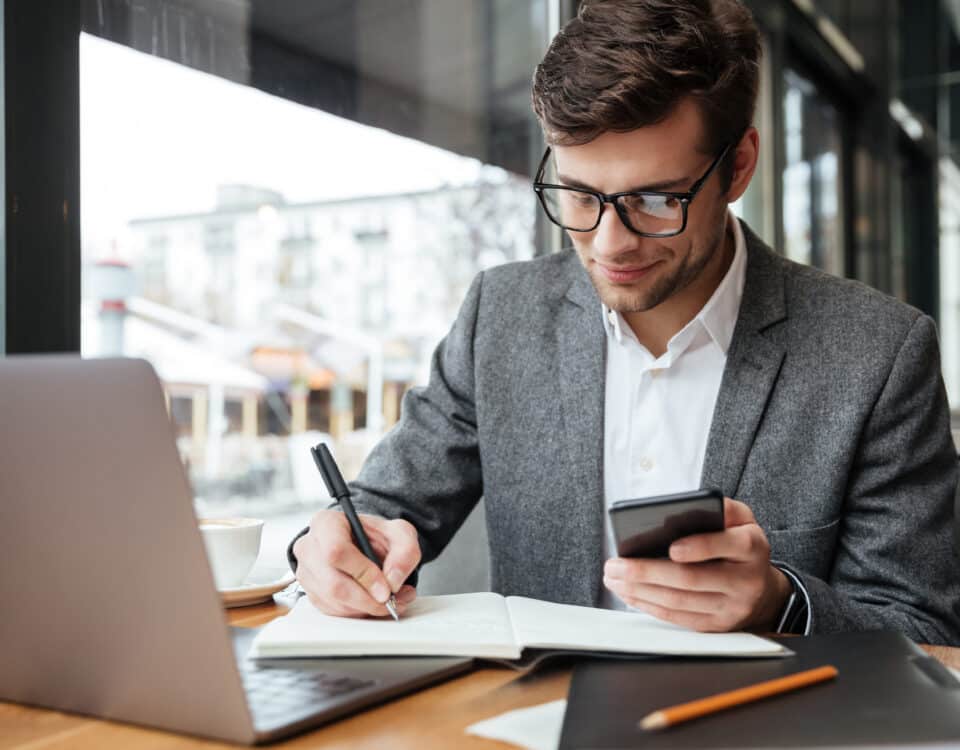 Um homem de óculos e terno senta-se em uma mesa com um laptop, concentrado em Registros SPED Contribuições, segurando um smartphone em uma das mãos e escrevendo em um caderno com a outra, totalmente envolvido em seu trabalho.