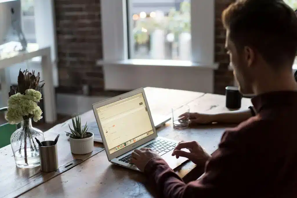 Uma pessoa está sentada a uma mesa de madeira digitando em um laptop, planejando uma plataforma omnicanal. Um copo d'água, dois pequenos vasos de plantas e materiais de escritório repousam sobre a mesa, enquanto a luz do sol entra pelas janelas ao fundo.