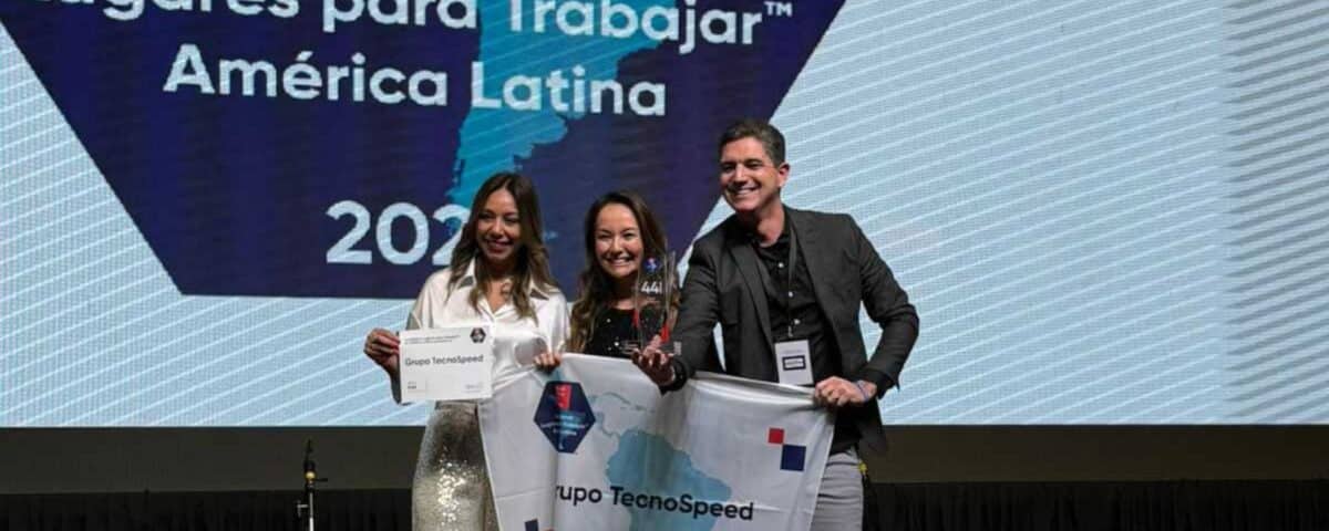 Três pessoas estão em pé no palco, sorrindo e segurando um certificado e uma bandeira com os dizeres "Grupo TecnoSpeed". Atrás delas, há uma tela grande exibindo o texto em espanhol: "Los Mejores Lugares para Trabajar América Latina 2022".