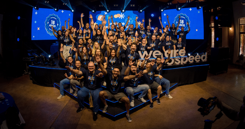 Descrição de Imagem: Um grande grupo de colaboradores da TecnoSpeed, vestindo camisas combinando, posam animadamente em um palco com o texto "18 anos" e "nós somos TecnoSpeed" ao lado de um logotipo do GPTW Brasil exibido em telas. O cenário é festivo, com iluminação brilhante e decorações realçando a atmosfera comemorativa.