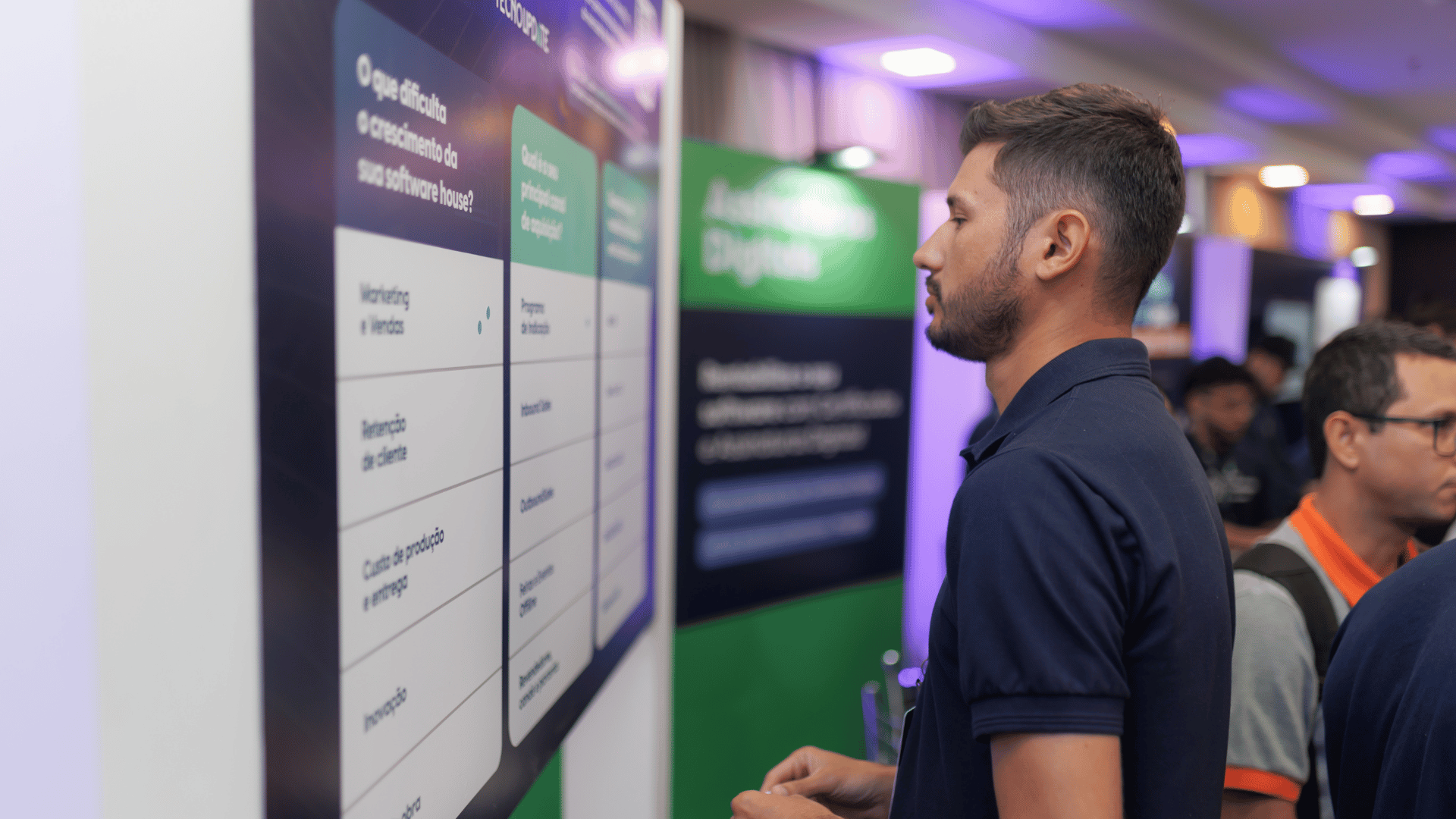 Um homem está em frente a um quadro informativo em um evento de tecnologia, profundamente absorto. A agitação da feira de negócios é evidente com faixas borradas e participantes promovendo uma atmosfera dinâmica de networking.