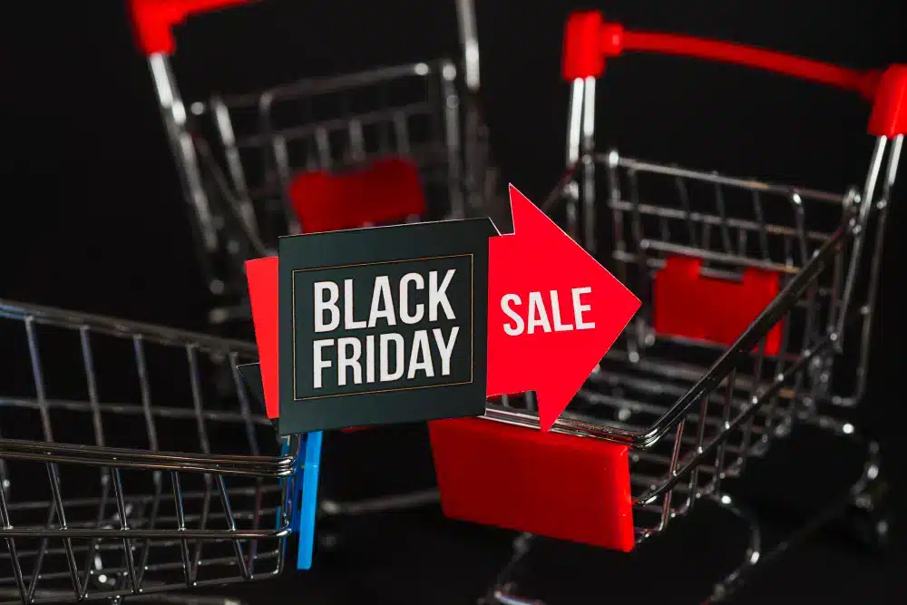 Vários carrinhos de compras em miniatura estão dispostos em um fundo preto, com um sinal de "Black Friday" e uma seta vermelha de "Sale" na frente, simbolizando as empolgantes ofertas de compras da Black Friday 2025.