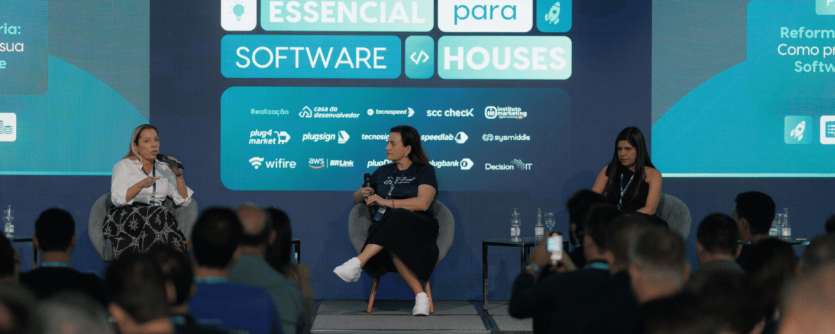 Um painel de três mulheres sentadas em um palco se envolvem em uma discussão no evento Technoupdate 15th Edition, aprofundando-se em tópicos como reforma tributária. O cenário apresenta um display digital com o tema “Essencial para Software Houses”. Uma plateia assiste atentamente.