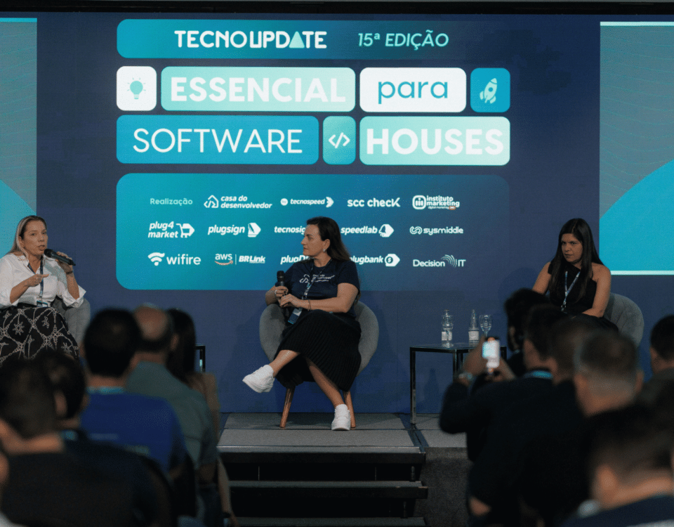 Um painel de três mulheres sentadas em um palco se envolvem em uma discussão no evento Technoupdate 15th Edition, aprofundando-se em tópicos como reforma tributária. O cenário apresenta um display digital com o tema “Essencial para Software Houses”. Uma plateia assiste atentamente.