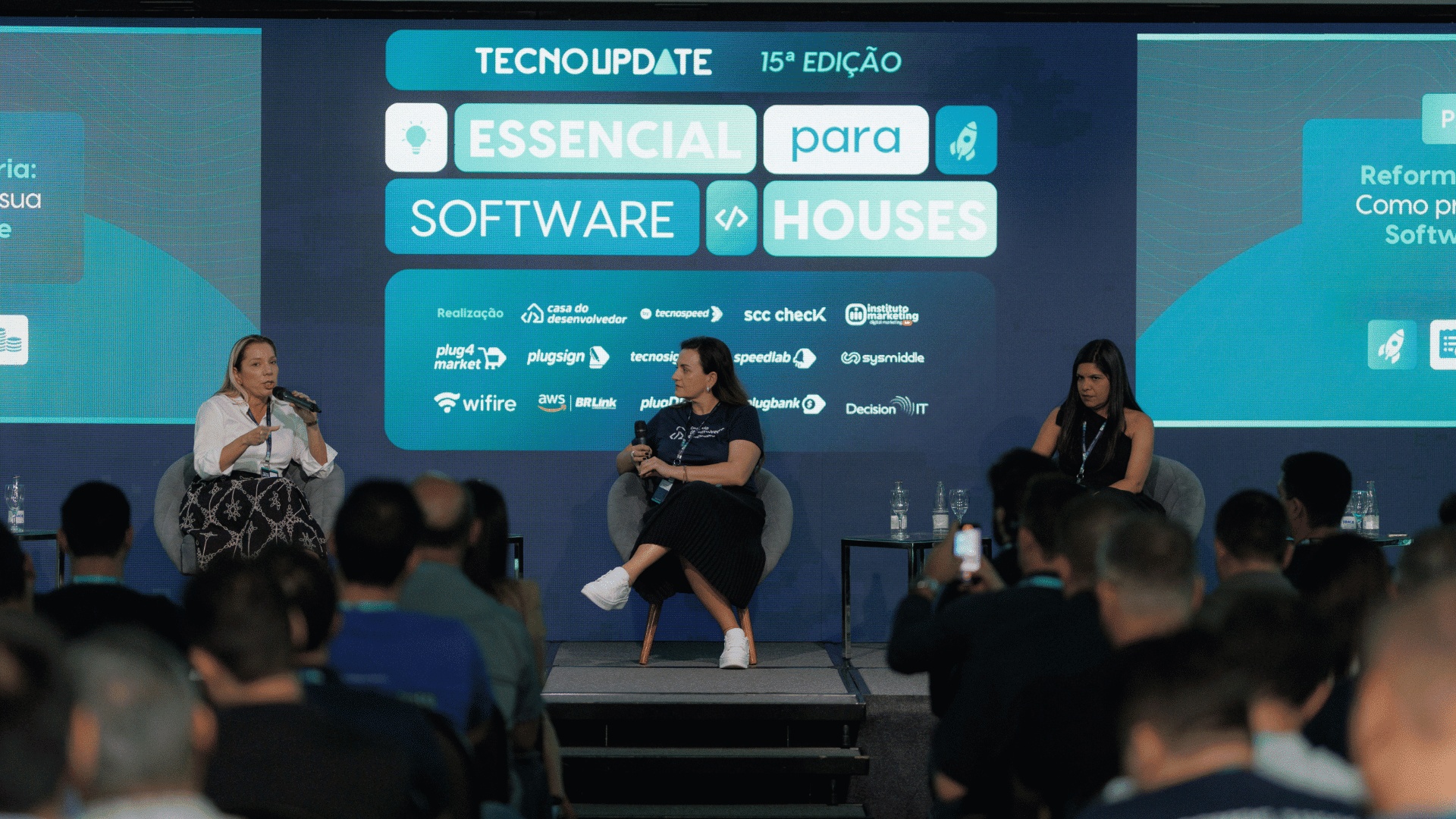 Um painel de três mulheres sentadas em um palco se envolvem em uma discussão no evento Technoupdate 15th Edition, aprofundando-se em tópicos como reforma tributária. O cenário apresenta um display digital com o tema “Essencial para Software Houses”. Uma plateia assiste atentamente.