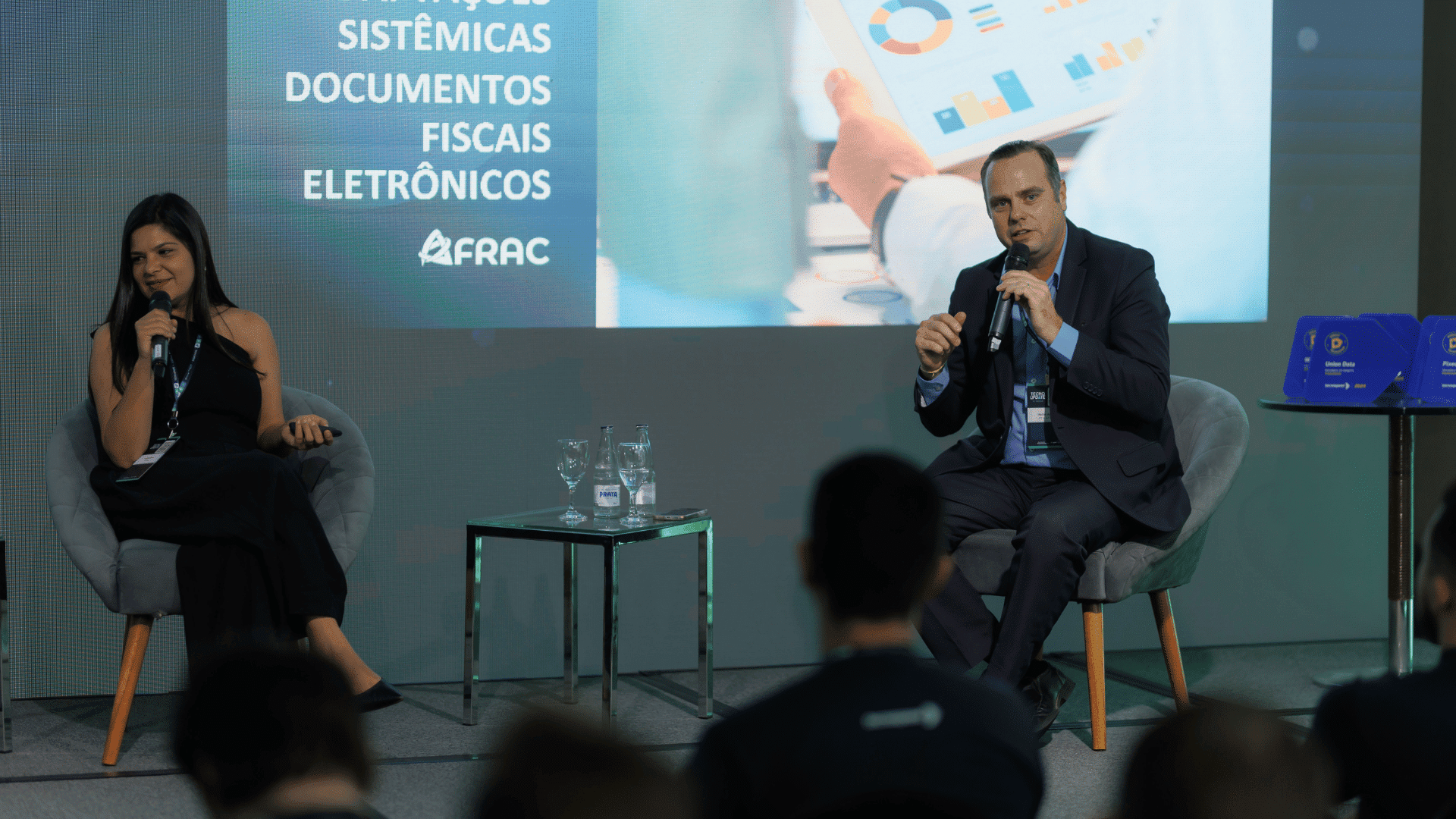 Dois indivíduos estão sentados no palco com microfones, envolvidos em uma discussão sobre reforma tributária. Uma tela atrás deles exibe gráficos e texto relacionado à tecnologia. Eles estão em um ambiente de conferência, com uma pequena audiência visível em primeiro plano.