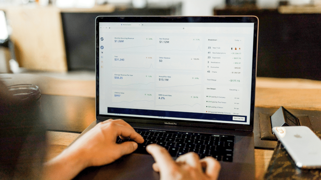 Uma pessoa usa um laptop exibindo análises financeiras com gráficos, números e indicadores de crescimento alimentados por uma API financeira. A tela mostra várias métricas e insights financeiros. Perto dali, um smartphone e um notebook repousam sobre a mesa de madeira.