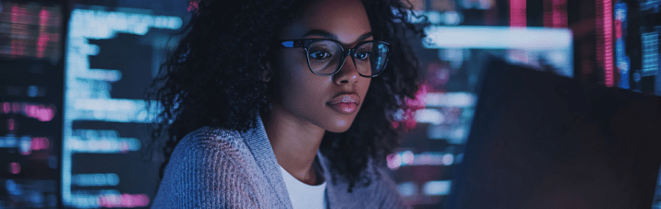 Descrição de Imagem: Uma mulher de óculos trabalha intensamente em um laptop, cercada por várias telas exibindo códigos coloridos. A sala mal iluminada exala uma atmosfera focada e voltada para a tecnologia, refletindo sua expertise em diversas áreas em TI.