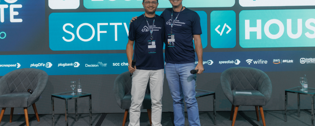 Dois homens estão no palco, sorrindo em camisetas combinando do evento. Atrás deles, uma tela exibe "Essencial Software" ao lado de vários logotipos. Como o novo CEO da TecnoSpeed, um homem exala confiança com duas cadeiras vazias complementando a configuração.