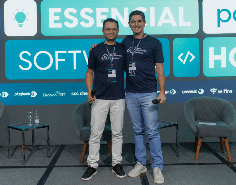 Dois homens estão no palco, sorrindo em camisetas combinando do evento. Atrás deles, uma tela exibe "Essencial Software" ao lado de vários logotipos. Como o novo CEO da TecnoSpeed, um homem exala confiança com duas cadeiras vazias complementando a configuração.