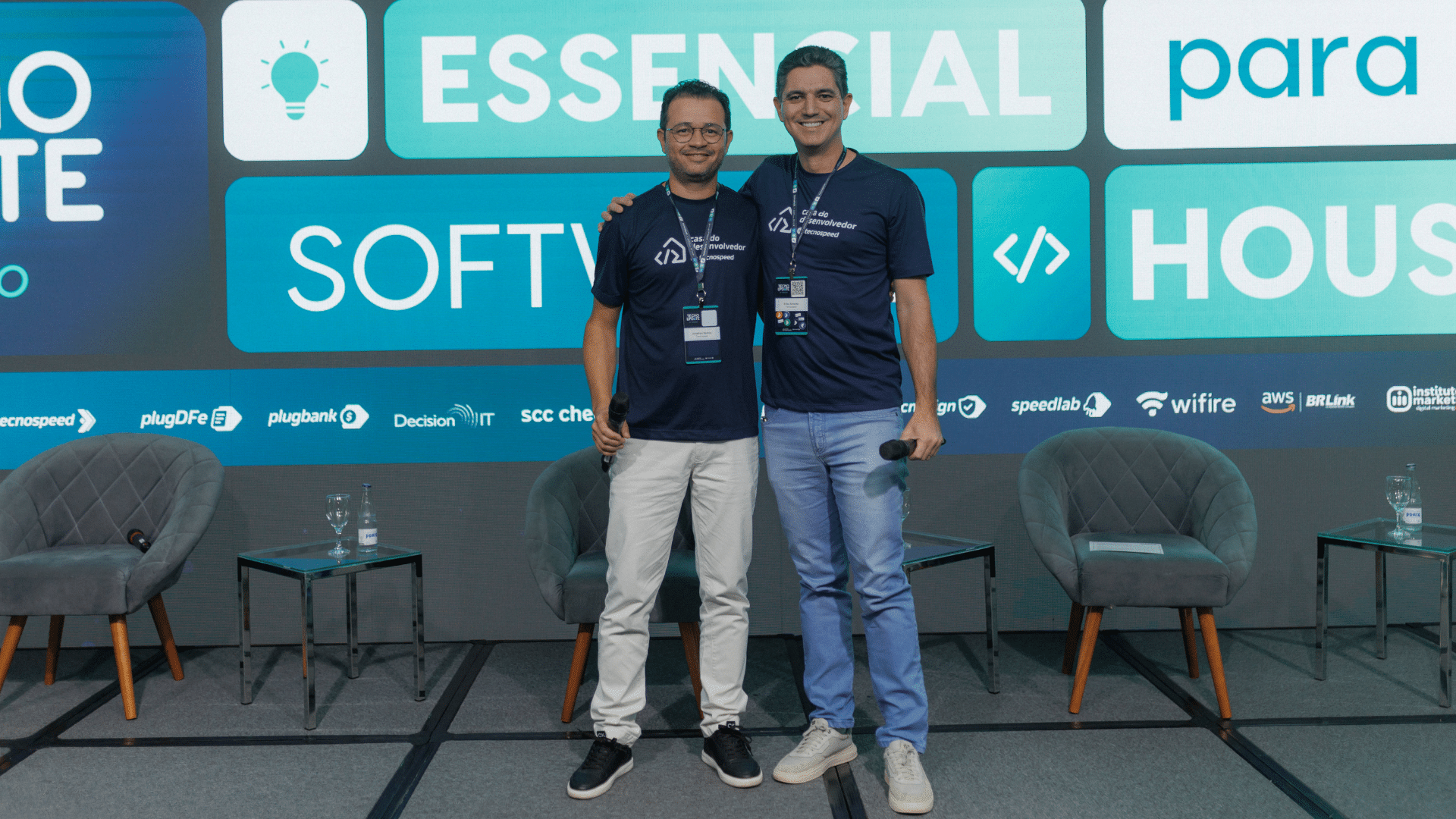 Dois homens estão no palco, sorrindo em camisetas combinando do evento. Atrás deles, uma tela exibe "Essencial Software" ao lado de vários logotipos. Como o novo CEO da TecnoSpeed, um homem exala confiança com duas cadeiras vazias complementando a configuração.