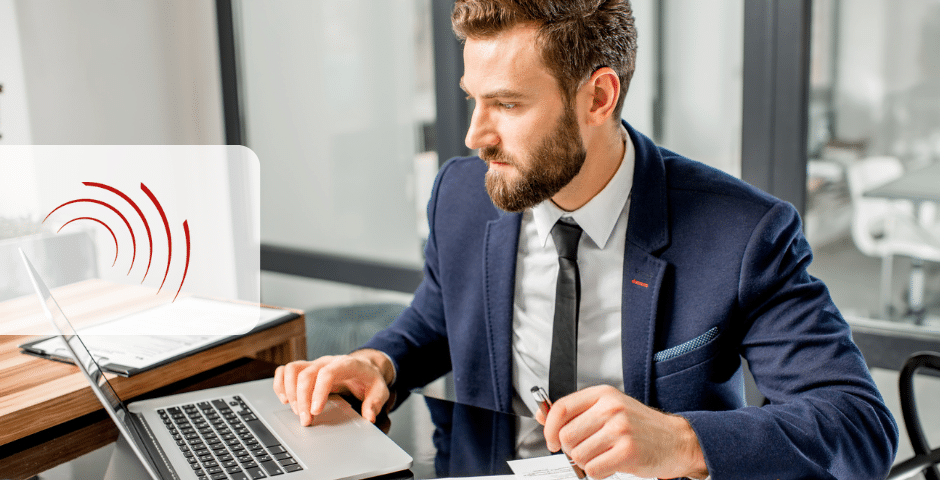 Um homem de terno está sentado em uma mesa, intensamente focado em seu laptop. Ele segura uma caneta e papel, possivelmente se aprofundando em Obrigações Acessórios no Brasil. O escritório moderno tem paredes de vidro e móveis elegantes, com um logotipo parcial visível à esquerda.