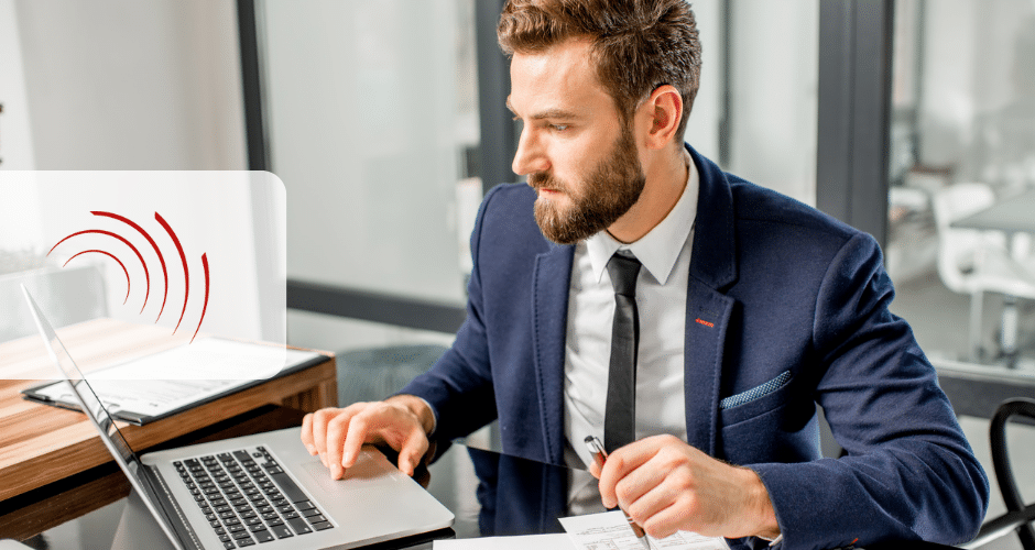 Um homem de terno está sentado em uma mesa, intensamente focado em seu laptop. Ele segura uma caneta e papel, possivelmente se aprofundando em Obrigações Acessórios no Brasil. O escritório moderno tem paredes de vidro e móveis elegantes, com um logotipo parcial visível à esquerda.