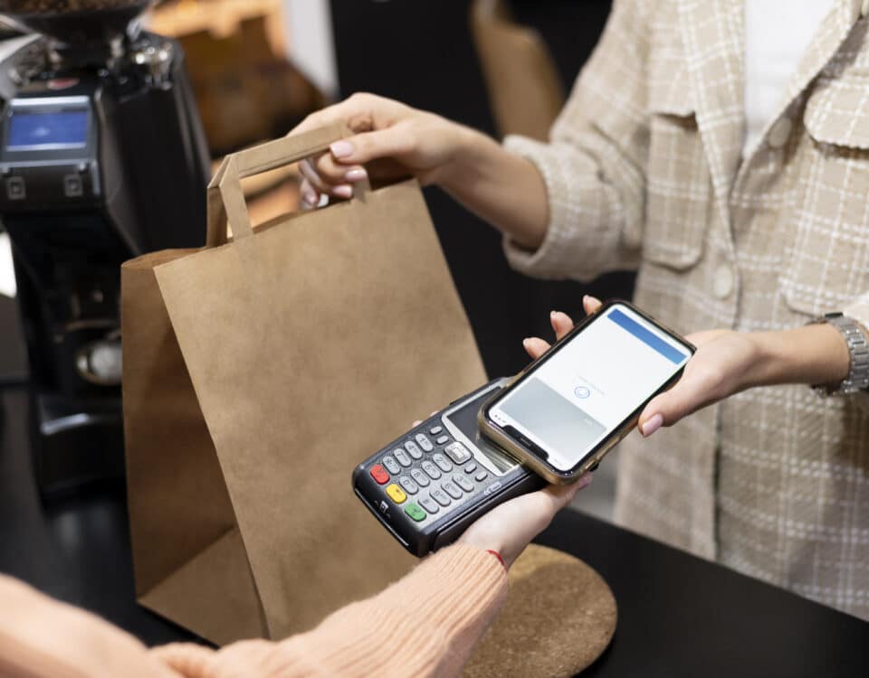 Uma pessoa segura uma sacola de papel pardo enquanto outra usa um smartphone, habilitado pelo sistema API NFC-e, para fazer um pagamento sem contato em um leitor de cartão em um balcão.