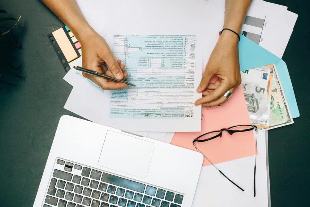 Uma pessoa preenche formulários de imposto de renda do Registro C100 SPED Fiscal em uma mesa com um laptop, óculos, dinheiro, documentos e post-its espalhados ao redor.