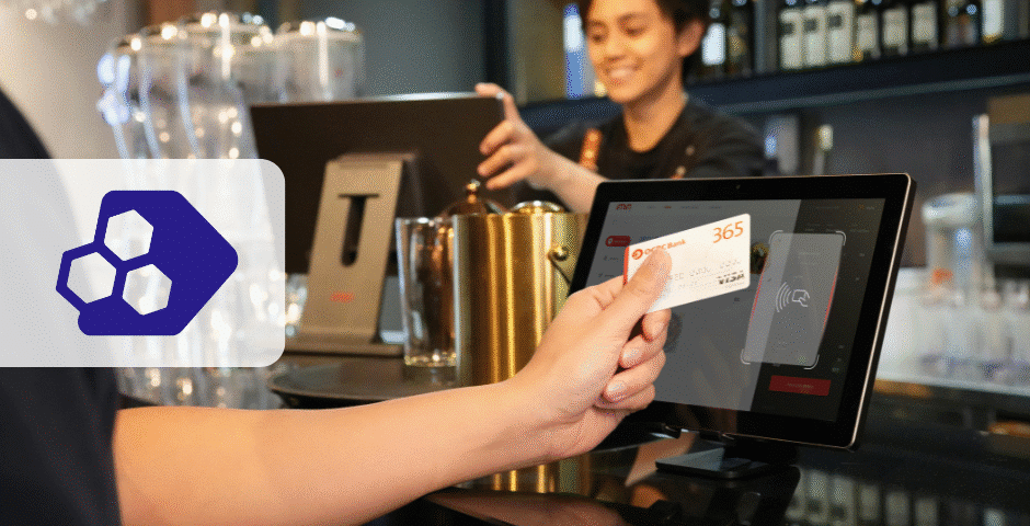Uma pessoa segura um cartão em um terminal de pagamento com tela sensível ao toque em um balcão, com um caixa sorridente ao fundo. À esquerda, aparece um logotipo geométrico azul, destacando a Reforma Tributária no Varejo no ambiente de varejo.
