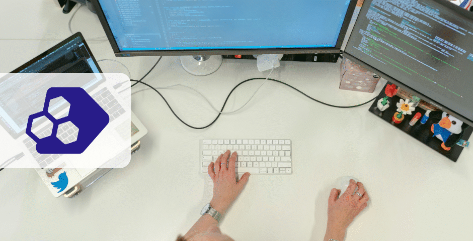 Uma pessoa está sentada em uma mesa branca usando um teclado e um mouse, com dois monitores exibindo códigos relacionados ao Desafios Reforma Tributária. Um laptop com um adesivo azul e algumas figuras pequenas também estão visíveis na mesa.
