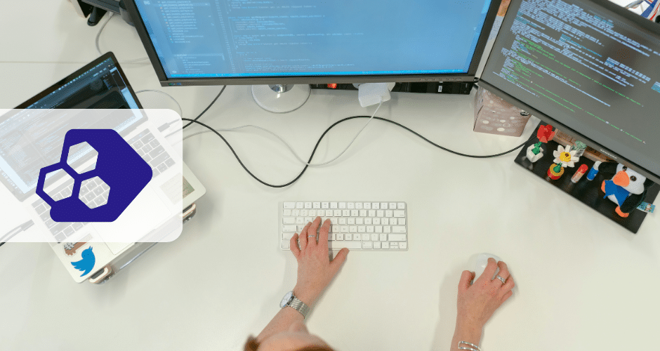 Uma pessoa está sentada em uma mesa branca usando um teclado e um mouse, com dois monitores exibindo códigos relacionados ao Desafios Reforma Tributária. Um laptop com um adesivo azul e algumas figuras pequenas também estão visíveis na mesa.