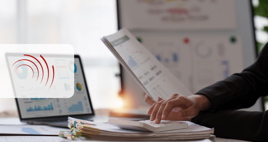 Uma pessoa usa uma calculadora enquanto segura documentos em uma mesa com gráficos financeiros em uma tela de laptop e papéis, sugerindo análise de dados ou implementação de reformas, como a reforma tributária do consumo. Um logotipo com linhas curvas vermelhas aparece à esquerda.