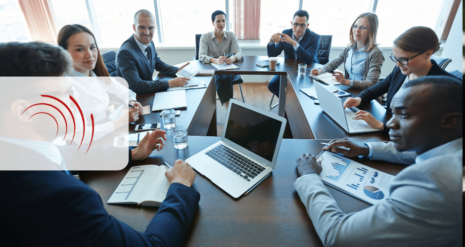 Um grupo de oito profissionais de negócios senta-se em torno de uma mesa de conferência com laptops, documentos e gráficos, discutindo benefícios fiscais em um escritório bem iluminado. Um ícone de som semitransparente sobrepõe parte da imagem.