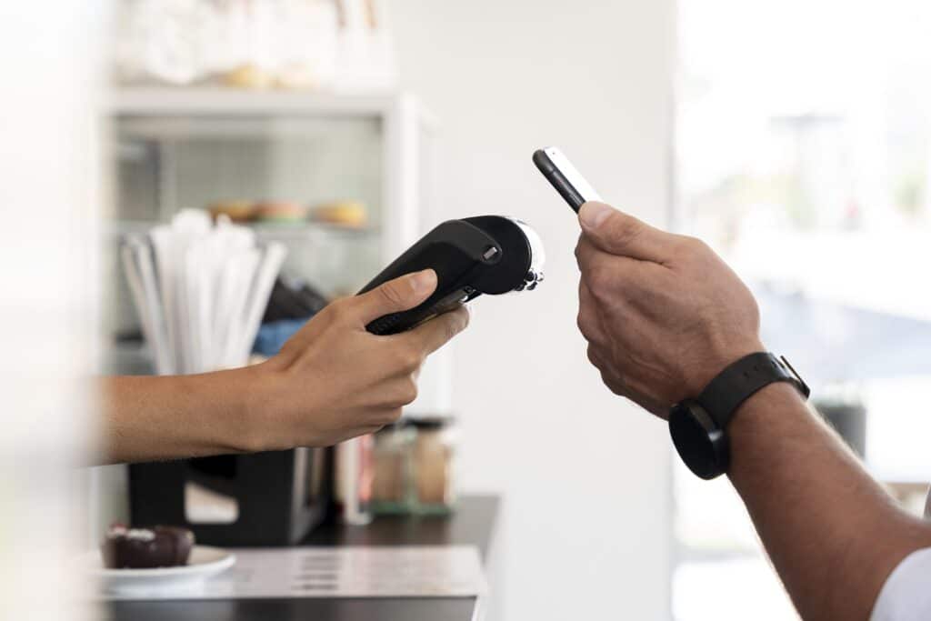 Uma pessoa segura um terminal de pagamento enquanto outra usa um smartphone para fazer um pagamento sem contato em um balcão, provavelmente preparando sua nota fiscal para varejo. Ambas as mãos estão visíveis; o plano de fundo mostra itens de cafeteria borrados.