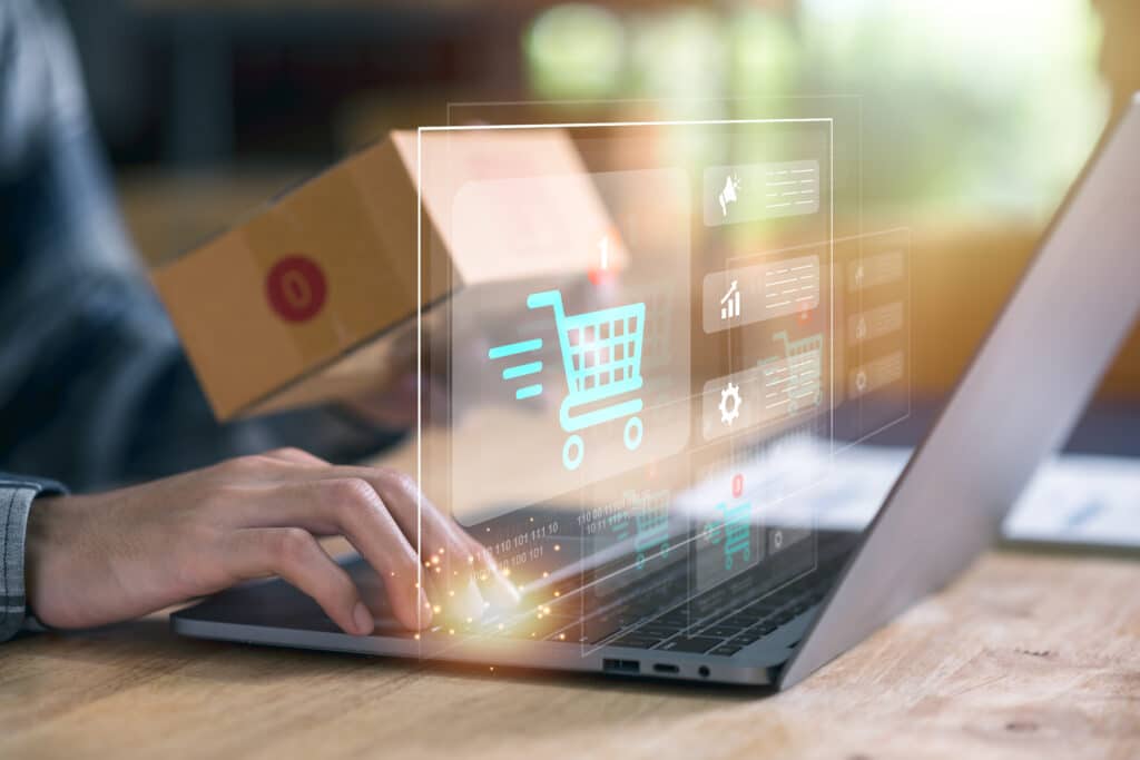 Uma pessoa usa um laptop com ícones digitais de comércio eletrônico, incluindo um símbolo de carrinho de compras, sobrepostos na tela - ilustrando uma gestão de marketplace. Uma mão digita no teclado enquanto a outra segura uma pequena caixa de papelão.