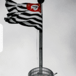 Uma bandeira listrada em preto e branco com um emblema vermelho tremula no topo de um mastro contra um céu nublado, fazendo referência a São Paulo. À esquerda, um ícone de documento roxo e branco da NFS-e de São Paulo se destaca na imagem.