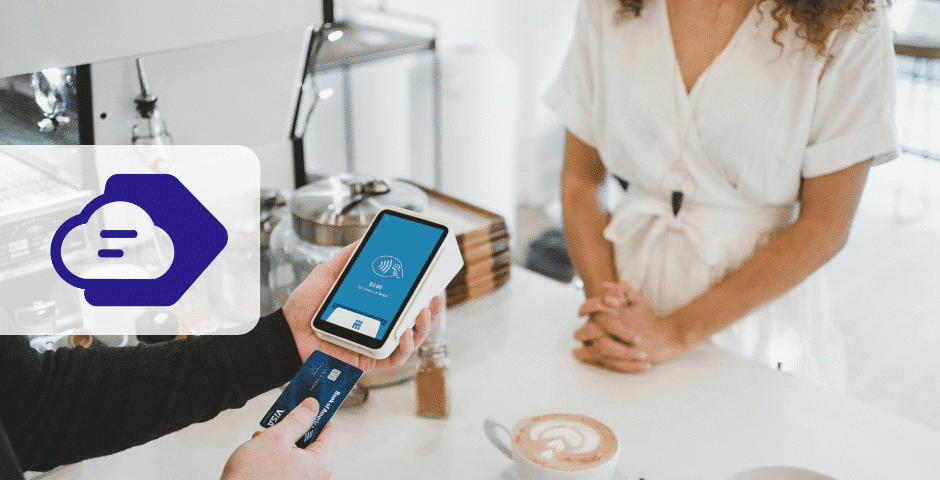 Uma pessoa segura um cartão de crédito e um smartphone exibindo um aplicativo bancário, demonstrando a vinculação dos meios de pagamentos enquanto se prepara para fazer um pagamento sem contato no balcão de uma cafeteria, com um barista em frente e uma xícara de café no balcão.