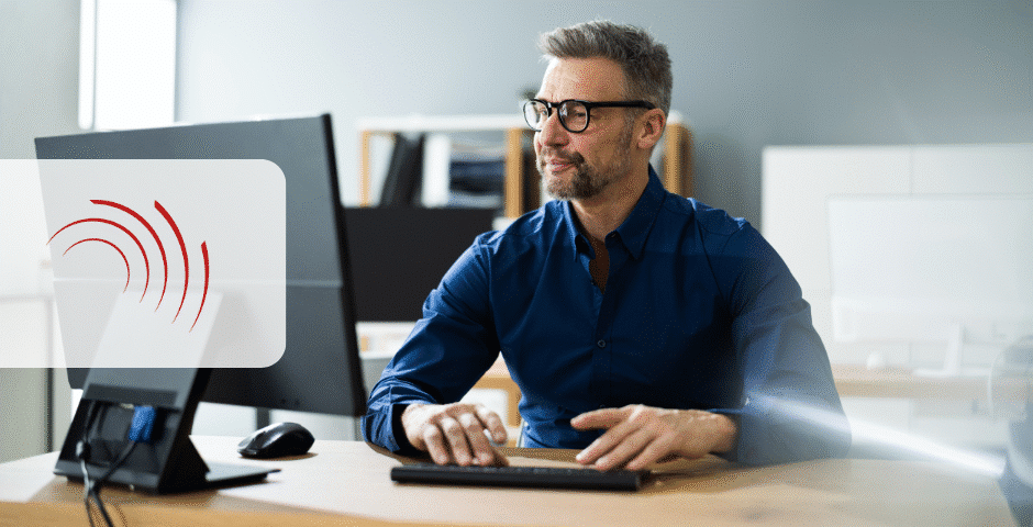 Um homem de óculos e cabelos grisalhos digita em um teclado em um escritório moderno, com foco em apuração assistida na reforma tributária. Um ícone de sinal digital vermelho é sobreposto à imagem.