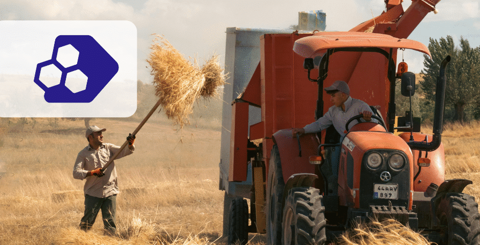 Dois homens colhem trigo em um campo; um deles usa um forcado para levantar a palha, enquanto o outro está sentado em um trator laranja. Um gráfico branco com hexágonos azuis no canto superior esquerdo destaca a Reforma Tributária para Produtor Rural.