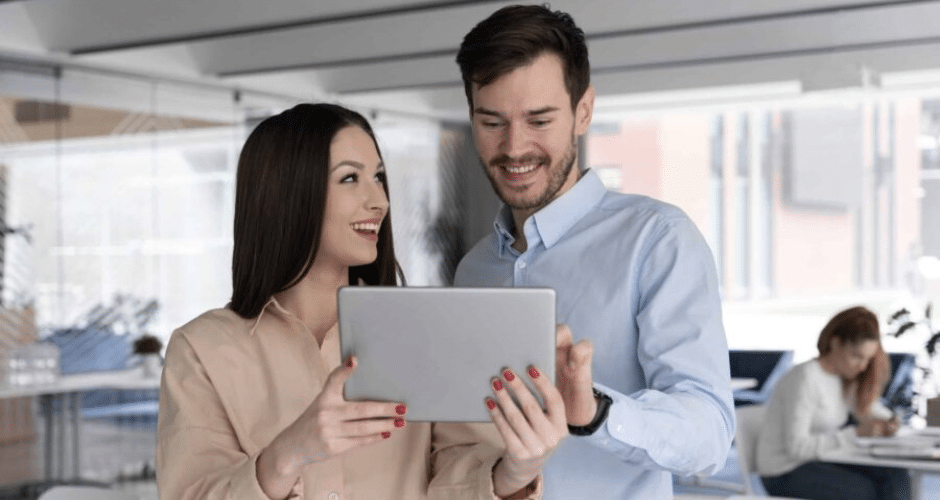 Dois colegas estão juntos em um escritório moderno, sorrindo e olhando para um tablet que a mulher está segurando, enquanto analisam a Vinculação de Pagamento NFCom em uma discussão de trabalho colaborativo.