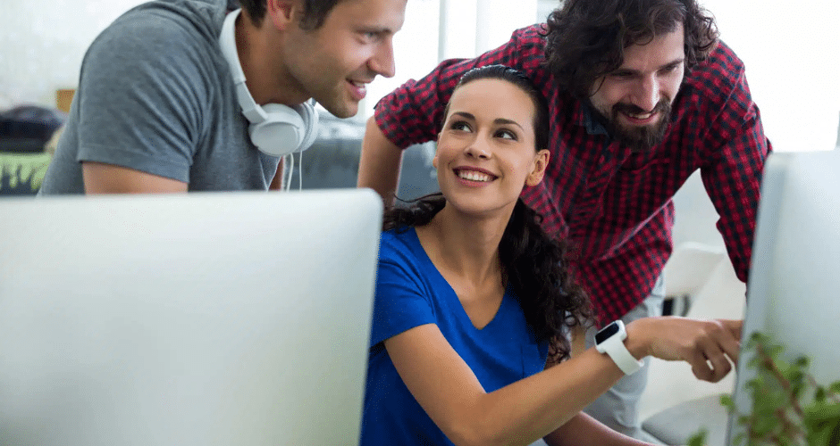 Três colegas estão reunidos em torno de uma mesa com computadores. Uma mulher está sentada, sorrindo e apontando para a tela, mostrando a Vinculação de Pagamento CT-e para dois homens ao seu lado, enquanto todos olham para o monitor e sorriem.