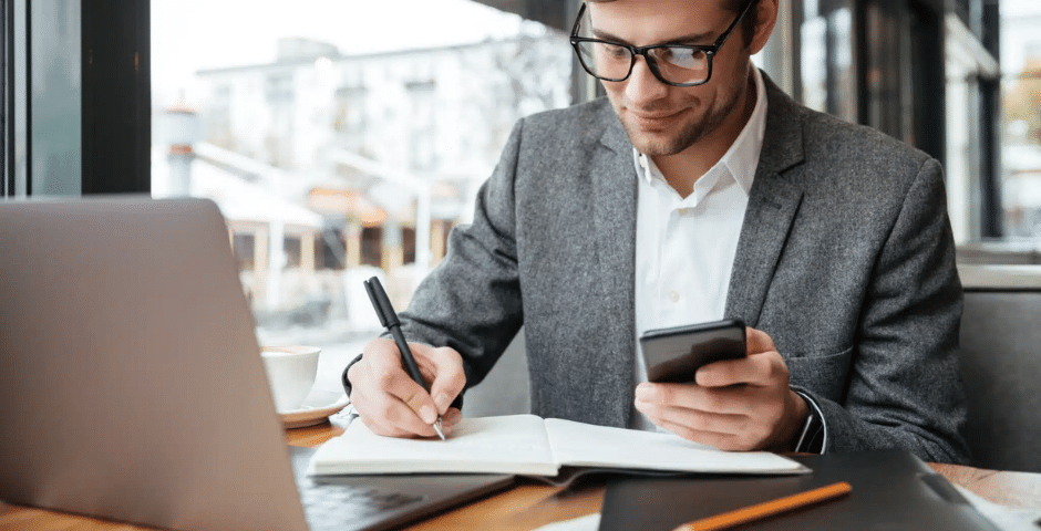 Um homem de terno senta-se em uma mesa com um laptop, um notebook e um telefone. Ele escreve no notebook enquanto segura o telefone, parecendo concentrado em questões como Descontinuidade da EFD-Contribuições. Grandes janelas revelam um cenário urbano.
