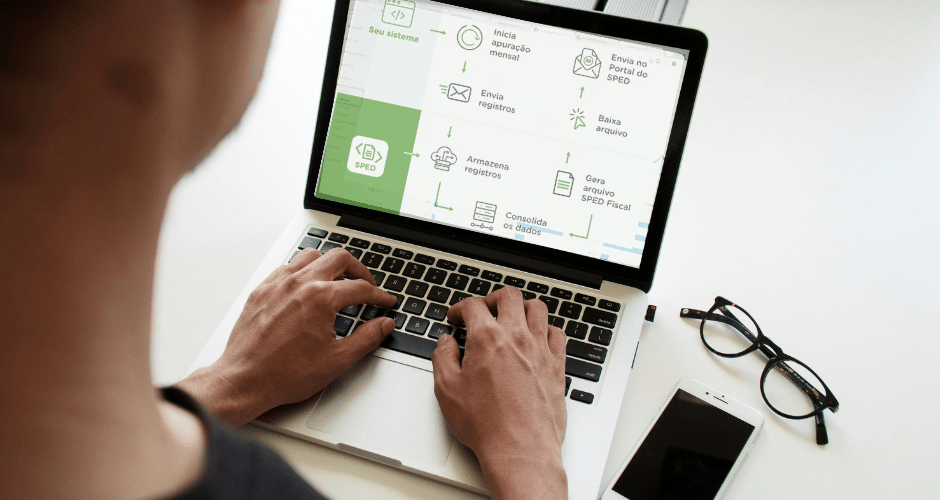 Uma pessoa digita em um laptop que exibe uma interface do software SPED XML com ícones verdes e texto em português. Ao lado, há óculos, um smartphone e uma caneta em uma mesa branca.