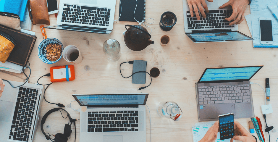 Uma visão de cima para baixo de uma mesa de madeira com cinco laptops abertos, várias mãos digitando sobre o SPED Automático, um smartphone, cadernos, lanches, fones de ouvido, um bule de chá e outros materiais de escritório espalhados.