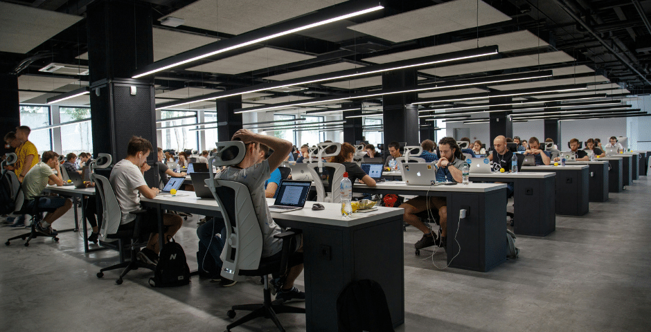 Grande escritório em plano aberto de uma empresa de software, com muitas pessoas sentadas em longas filas de mesas, trabalhando em laptops. As luzes brilhantes e as grandes janelas criam uma atmosfera moderna, movimentada e colaborativa no espaço de trabalho.