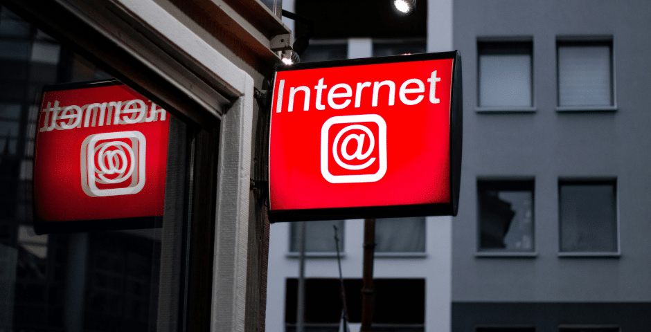 Uma placa vermelha com a palavra "Internet" e o símbolo @ está instalada em um prédio, refletindo em uma janela próxima - indicando a presença da Tributação sobre serviços de telecomunicações em meio ao cenário cinza de um apartamento ou escritório.